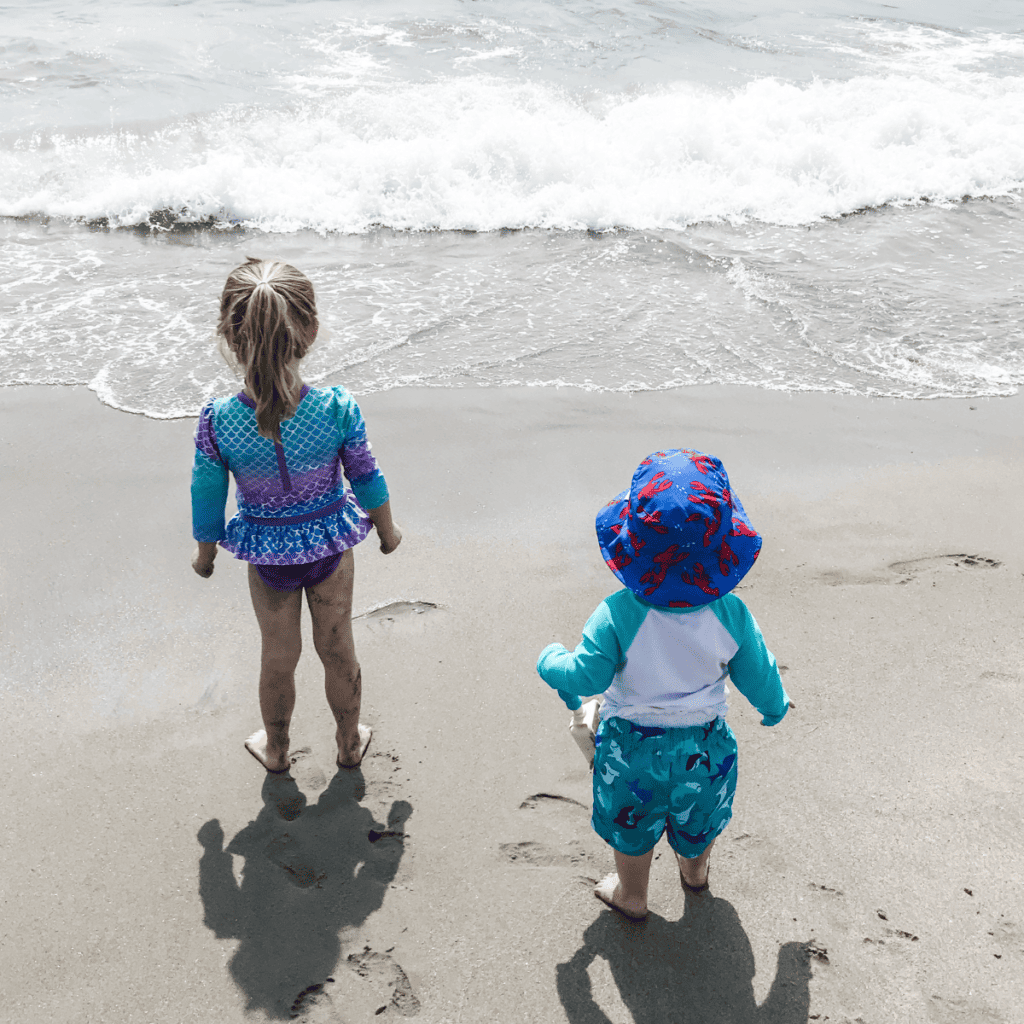 Photo of 2 children standing on the sand at the water's edge at the ocean.